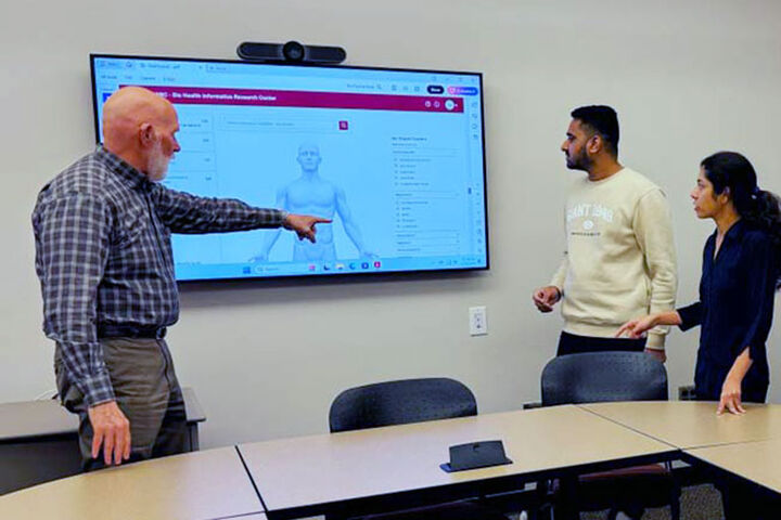 A male professor points to data on a large monitor while students look on.